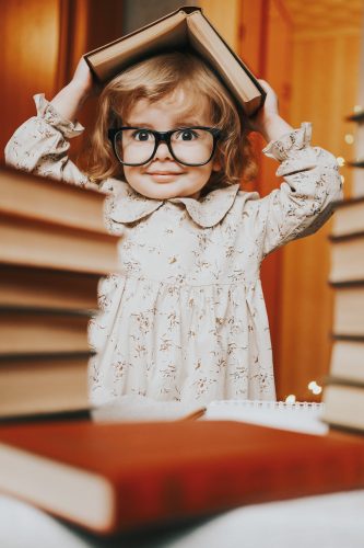 A little girl with glasses covered her head with a book. Cheerful child with book